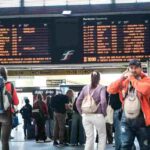 Persone attendono in stazione guardando il tabellone treni con cancellazioni e ritardi