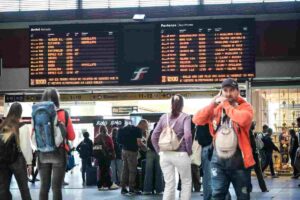 Persone attendono in stazione guardando il tabellone treni con cancellazioni e ritardi